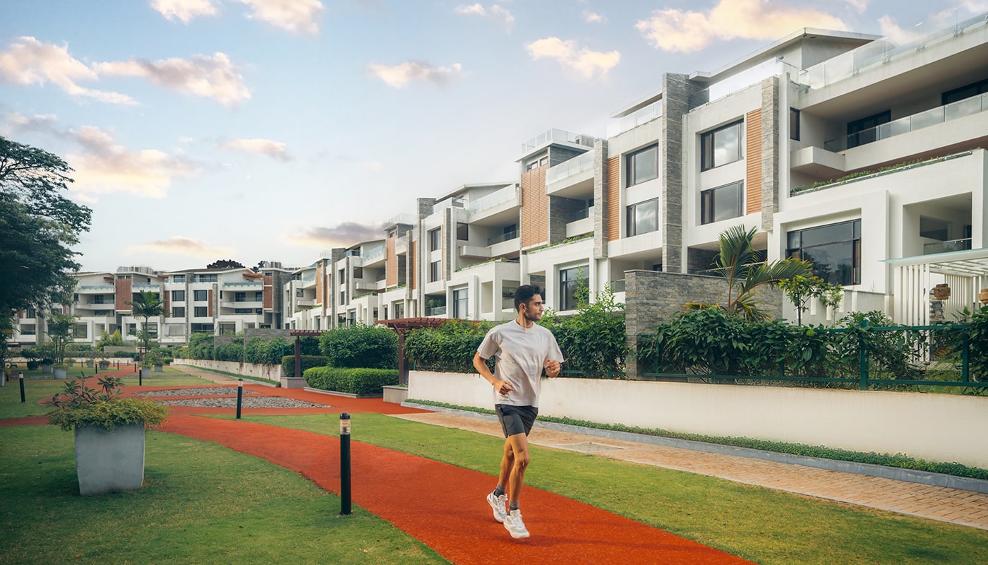 Man jogging on a red jogging track amidst lush greenery at Embassy Grove, with luxury villament residences in the backdrop under a serene evening sky in Bangalore