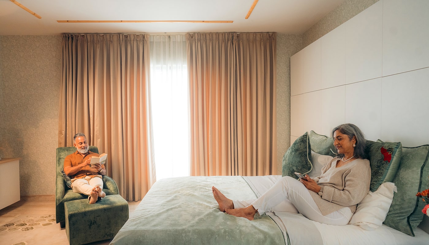 Parents enjoying a peaceful moment in a luxurious Embassy Grove bedroom – he reads in a plush armchair while she relaxes on the bed, reflecting serene and elegant living in Bangalore