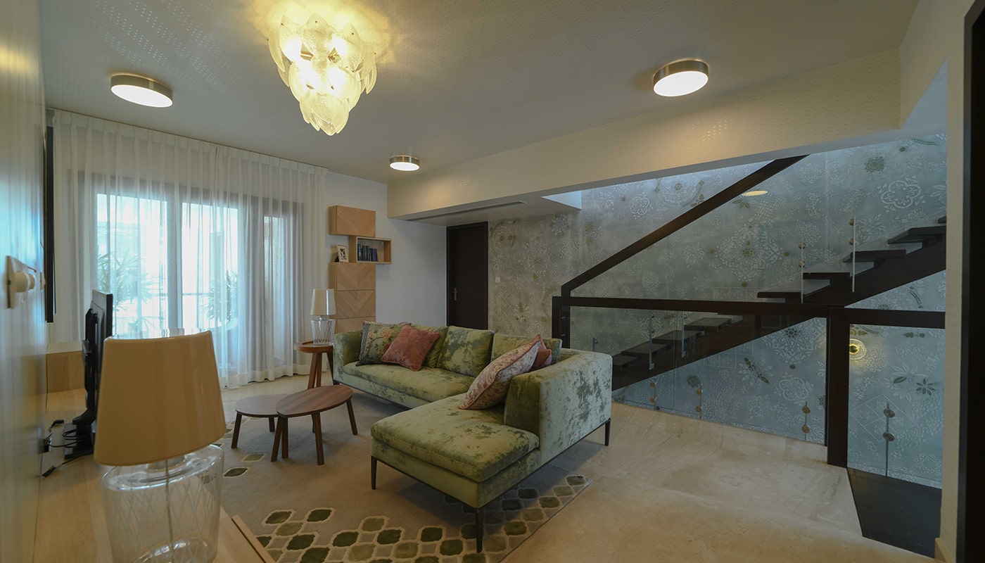 Modern lounge area in an Embassy Grove villament featuring a designer green velvet sectional sofa, artistic staircase with glass railing, ambient lighting, and elegant décor.