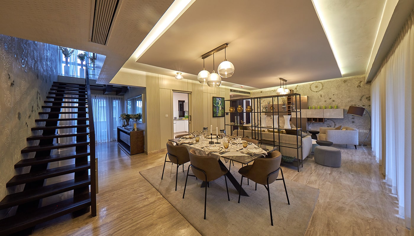 Luxurious open-plan dining and living space in an Embassy Grove villament featuring modern pendant lighting, designer dining setup, artistic decor, and a sleek floating staircase in Bangalore.